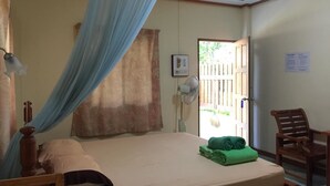 A bedroom with a bed, a chair, a bedside table, a fan, and a window with curtains. - Sandee Bungalows (Ko Pha-ngan)