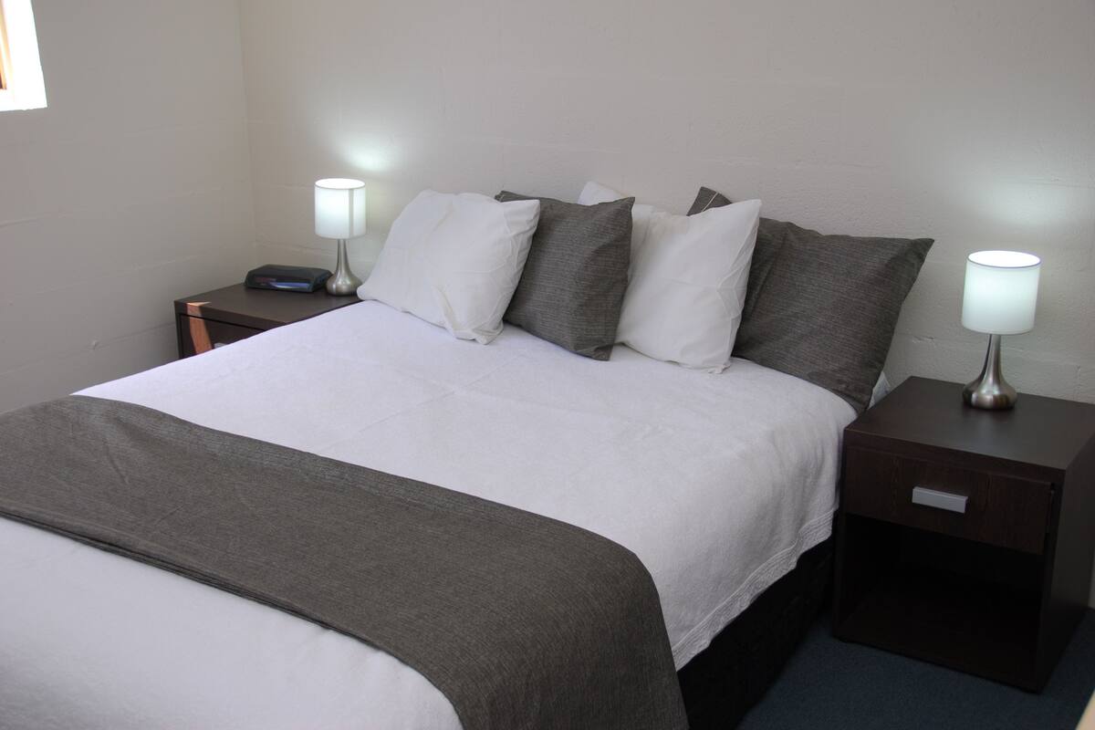 A neatly made bed with a white and gray bedspread, two bedside tables with lamps, and a dark nightstand.