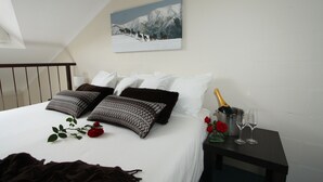 A neatly made bed with white linens and decorative pillows, a framed picture on the wall, and a bedside table with a bottle of champagne and glasses. - Summit Ridge Alpine Lodge (Falls Creek)