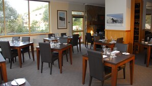 A dining area with multiple tables set for guests, large windows offering a view of the outdoors, and a fireplace with a mounted painting. - Summit Ridge Alpine Lodge (Falls Creek)