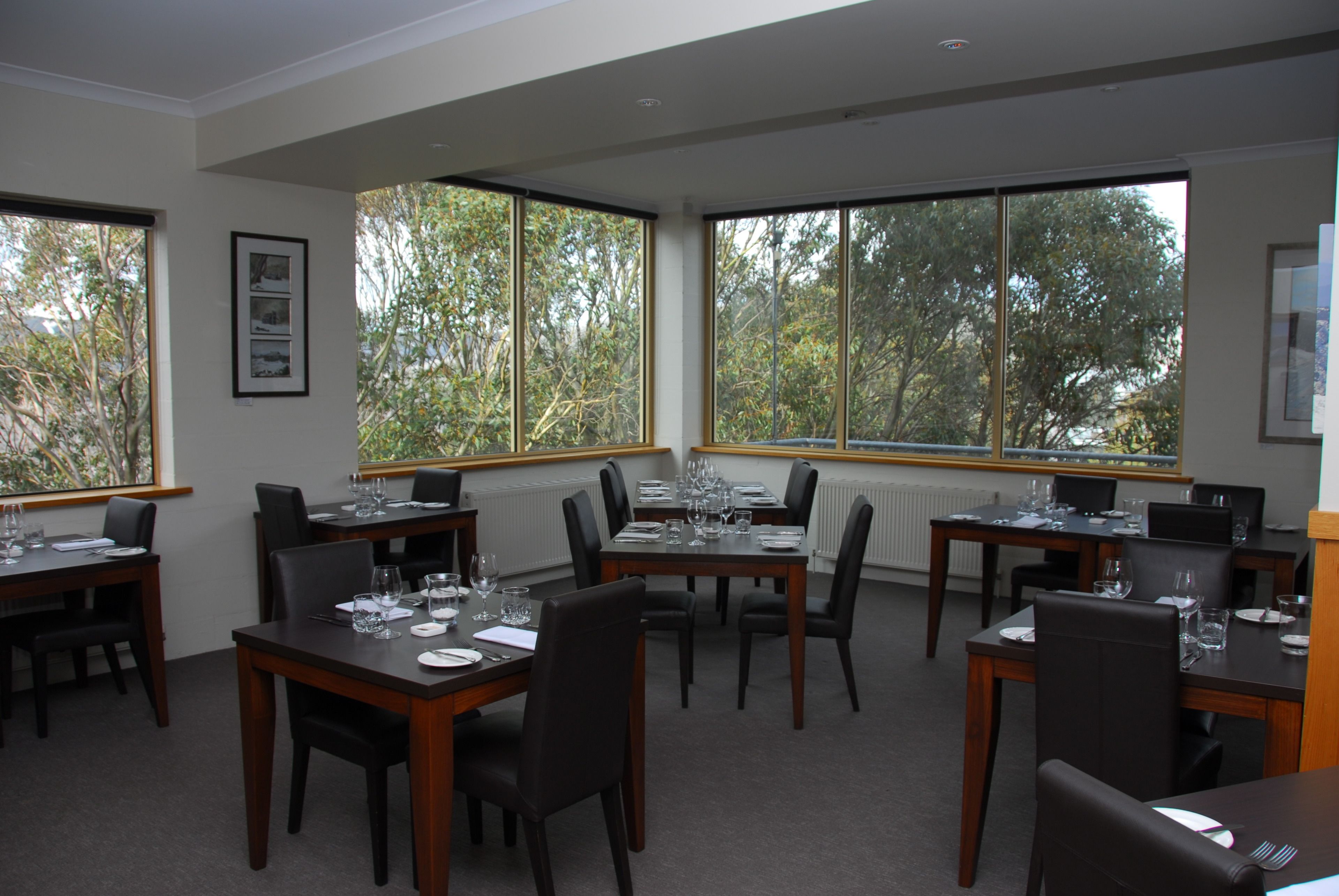 A dining area with multiple tables set for meals, large windows offering a view of trees, and framed artwork on the walls.