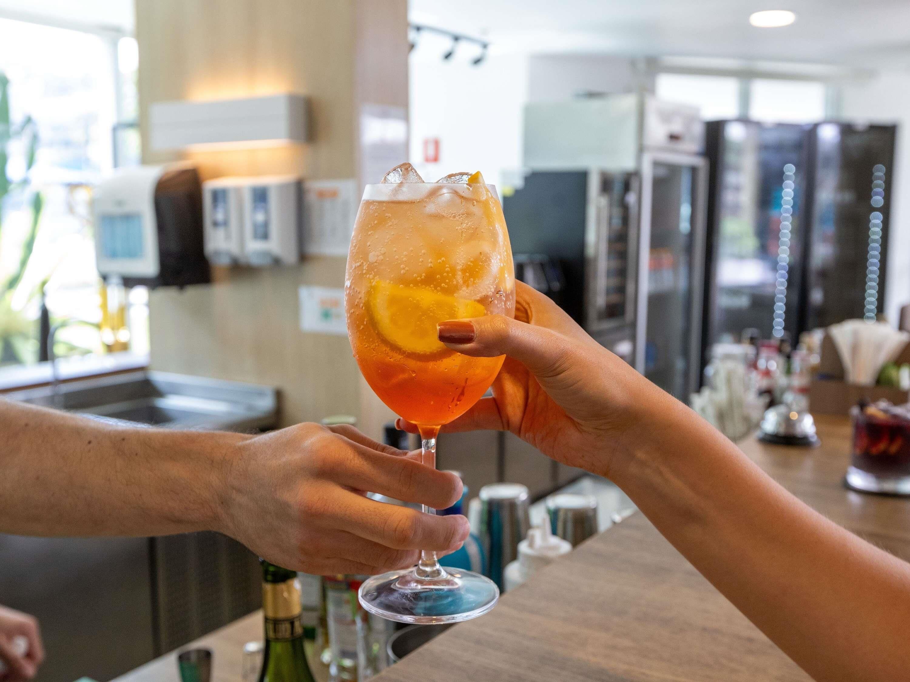 Deux mains tenant un verre dans lequel on a mis une tranche d’agrumes, une bouteille et un comptoir de bar, le tout dans un décor moderne.