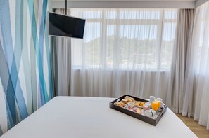 Ett hotellrum med en säng, ett bricka med frukosttillbehör, en platt-TV och tunna, genomskinliga gardiner.