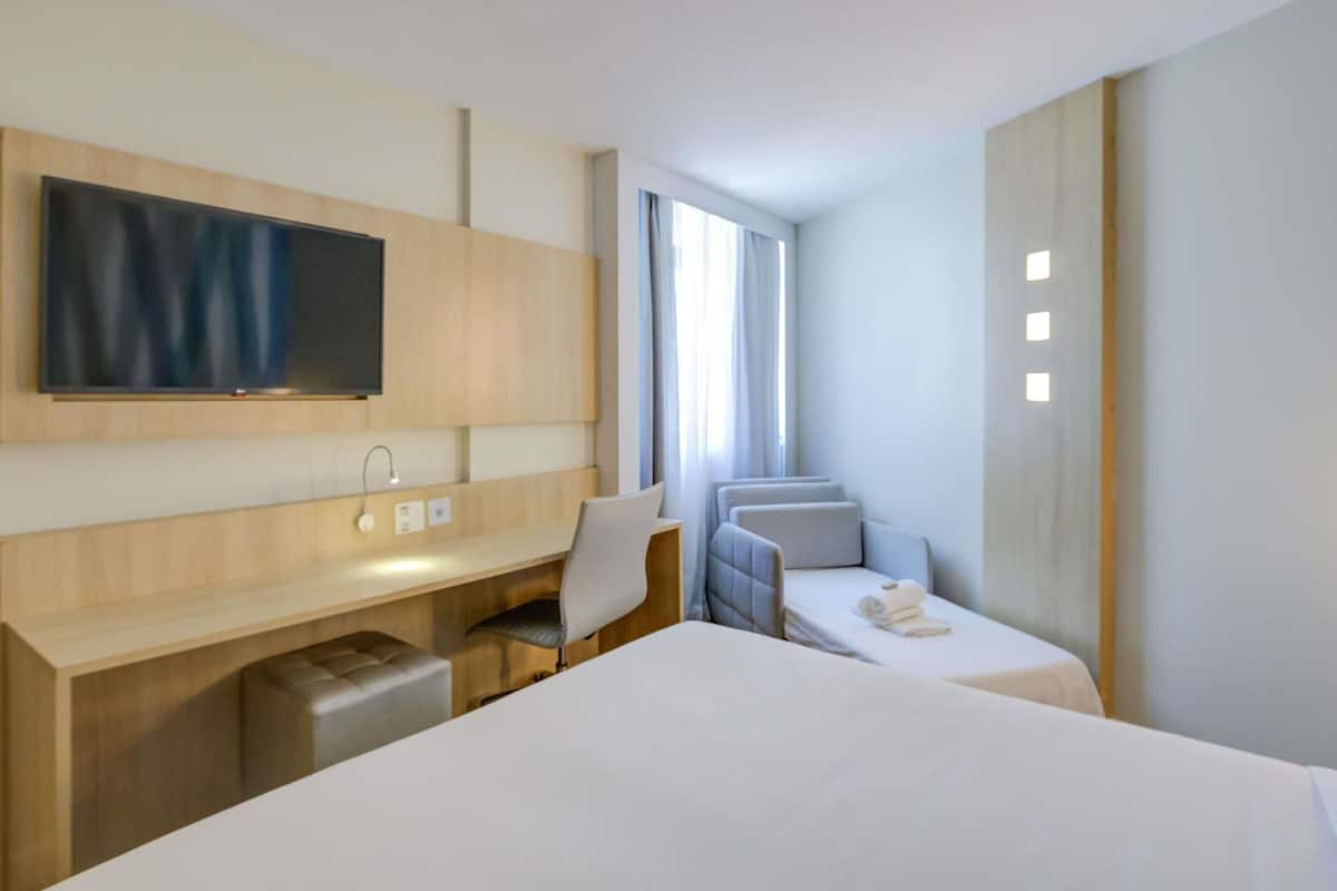 Moderni hotellihuone, jossa on suuri sänky, työpöytä tuolilla, seinälle asennettu televisio ja ikkuna, jossa on verhot.