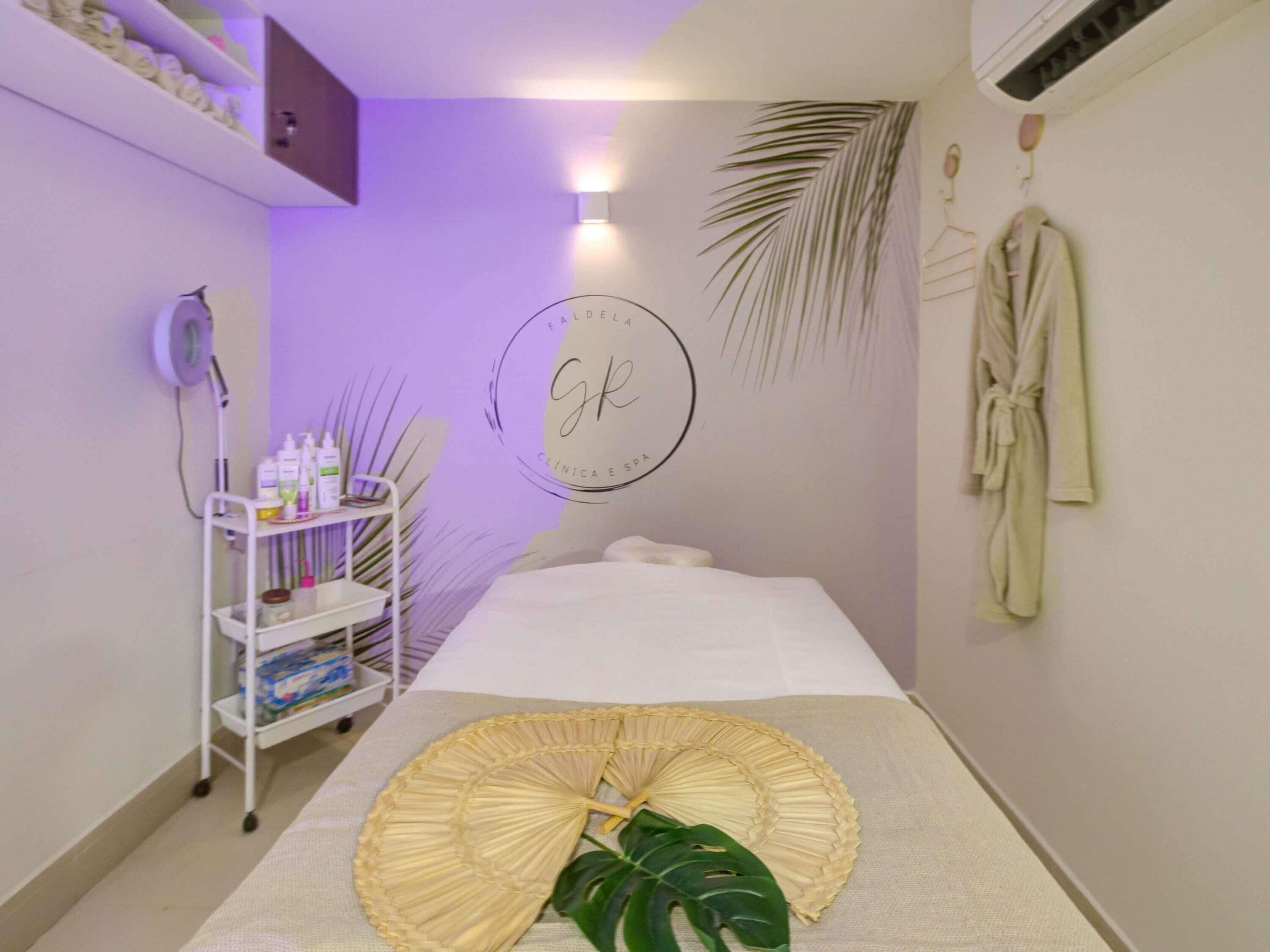 Um quarto de spa com mesa de massagem, prateleira com frascos, ventilador e parede decorada com folha de palmeira.