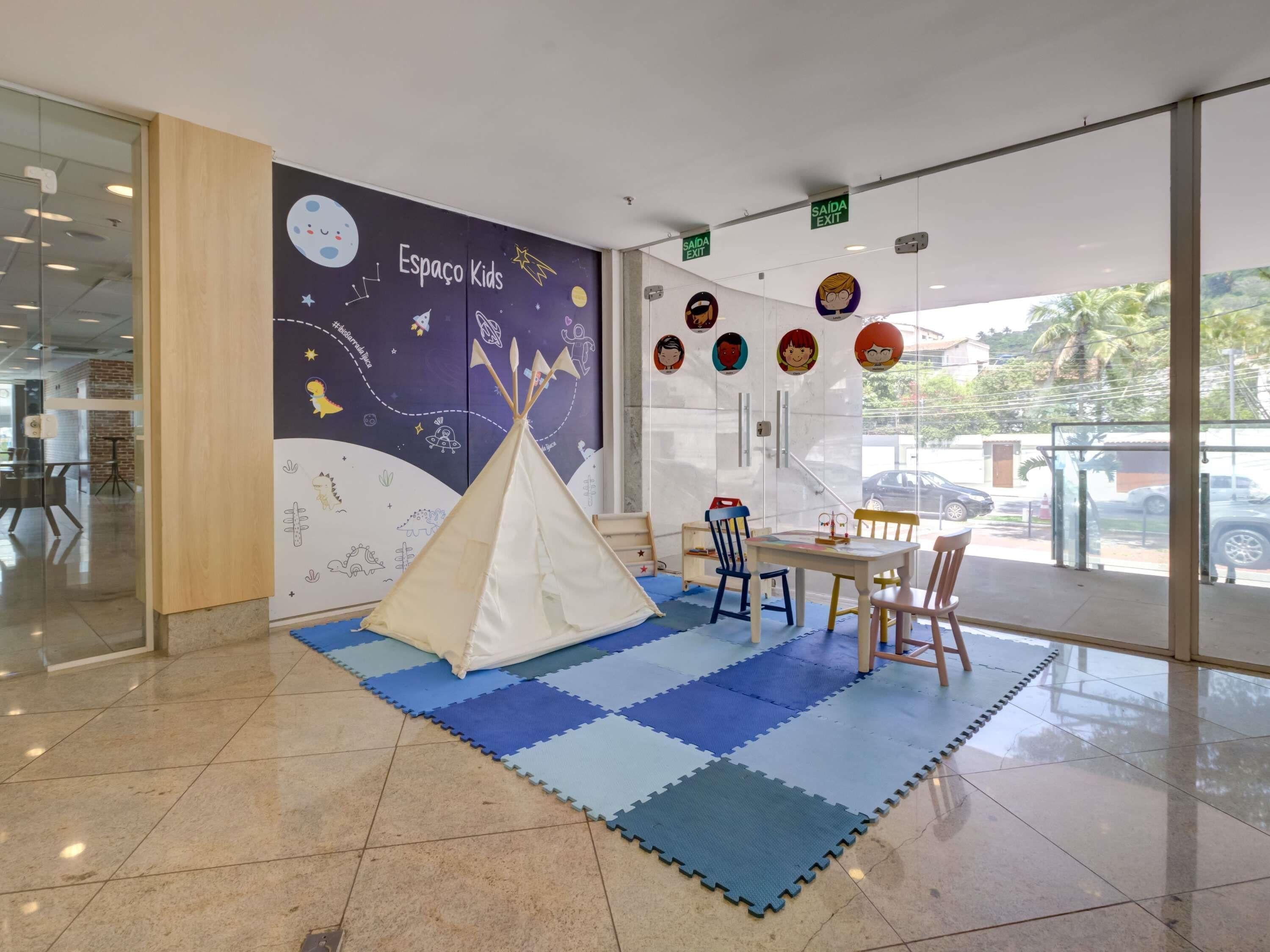 Uma área de recreação infantil com um mural com tema espacial, um tipi branco e uma mesa com cadeiras.