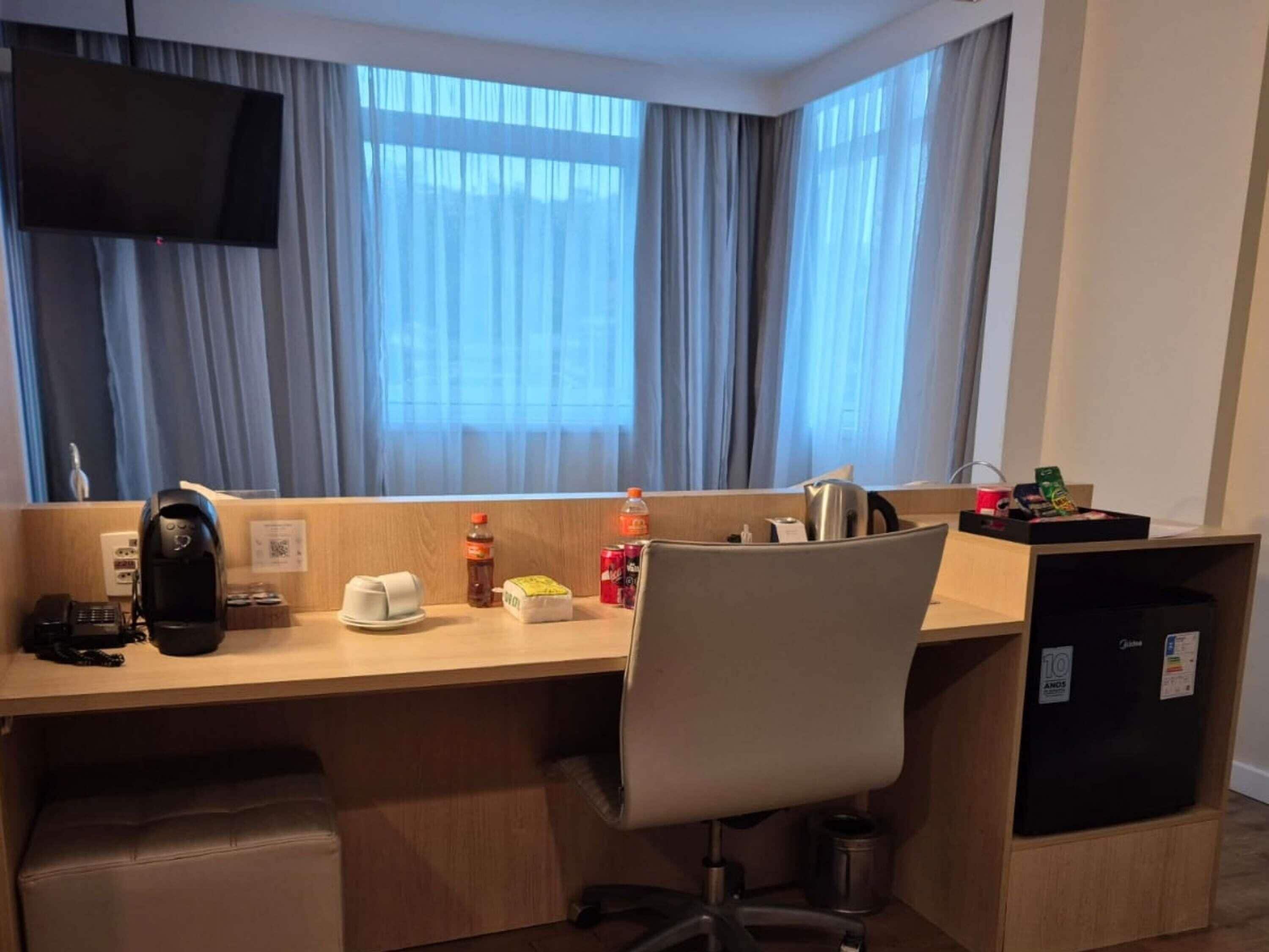 A hotel room with a desk, chair, coffee maker, kettle, and a refrigerator.