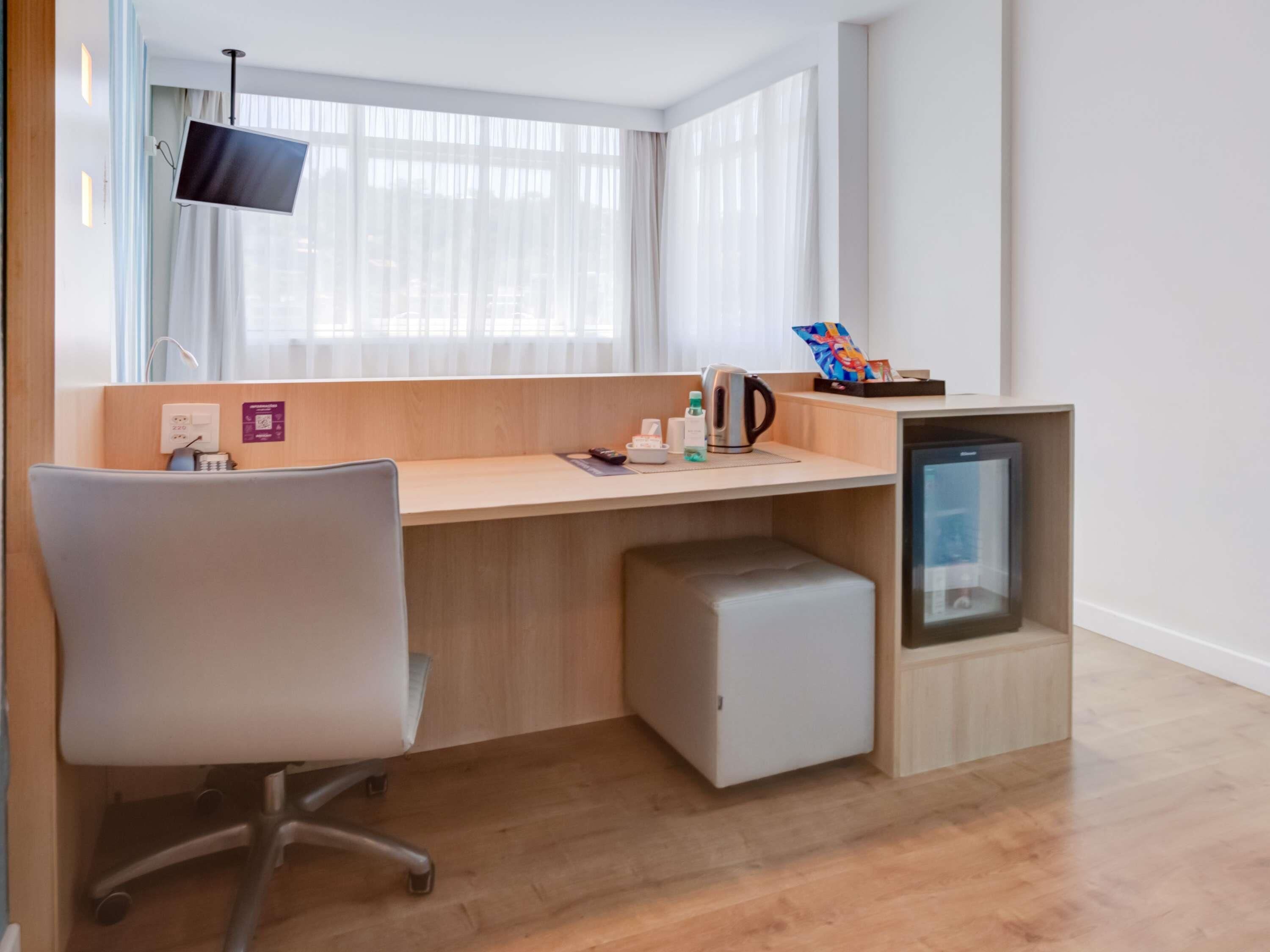 Une chambre d’hôtel moderne avec un bureau en bois, une chaise, un petit réfrigérateur, un téléviseur fixé au mur et une fenêtre avec des rideaux légers.
