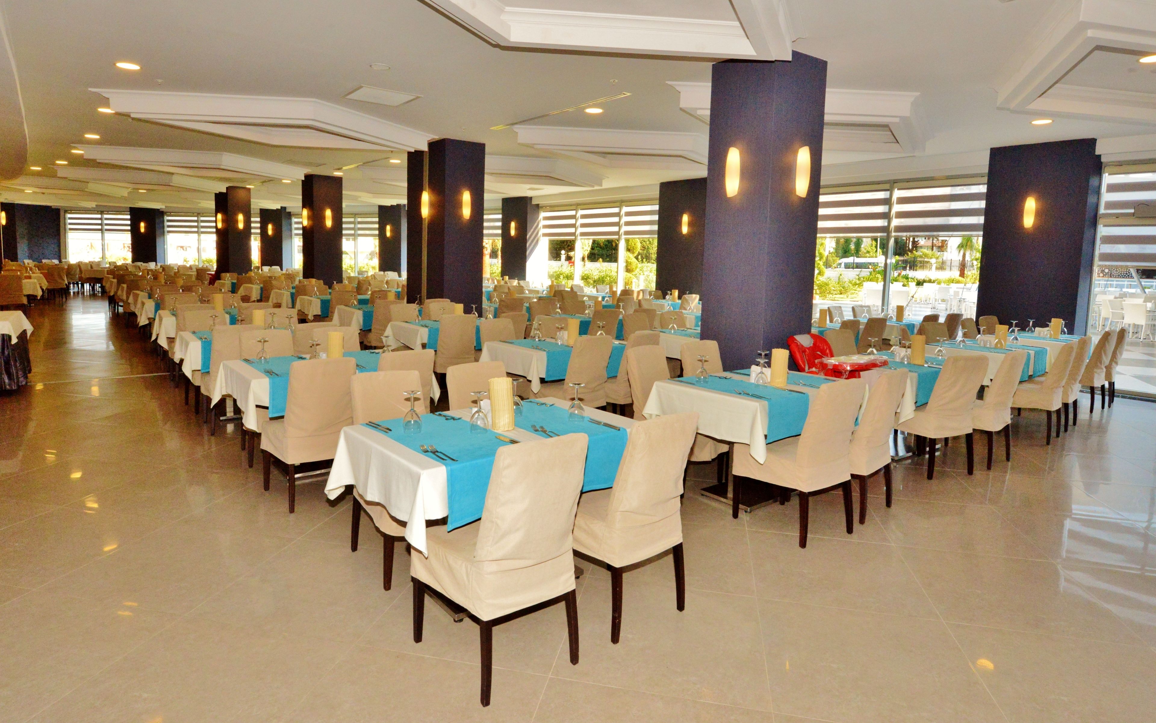 Un amplio salón de banquetes con filas de mesas, cada una con manteles blancos y centros de mesa azules, y altos pilares con luminarias.