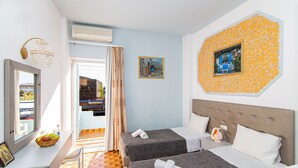 A hotel room with two beds, a window with a view, a wall-mounted air conditioner, and a decorative wall piece. - Minoa Hotel (Hersonissos)