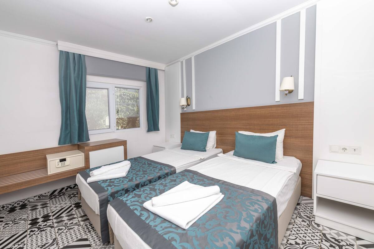 A hotel room with two beds, a window with curtains, a bedside table, a nightstand, and a patterned floor.