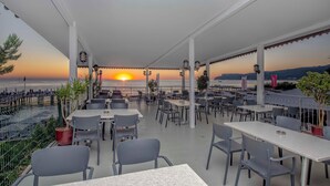 Un área de comedor al aire libre con mesas y sillas, con vista a una puesta de sol sobre el mar.