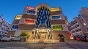 Un hotel de varios pisos con un atrio central, un llamativo letrero 'ARMA'S' y un estacionamiento con varios automóviles.