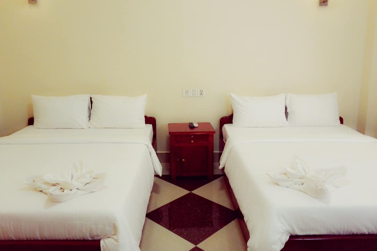 A hotel room with two beds, each with white linens and a decorative white towel. There is a small wooden nightstand between the beds with a remote control on it. The room has a simple, clean design with a checkered floor pattern.