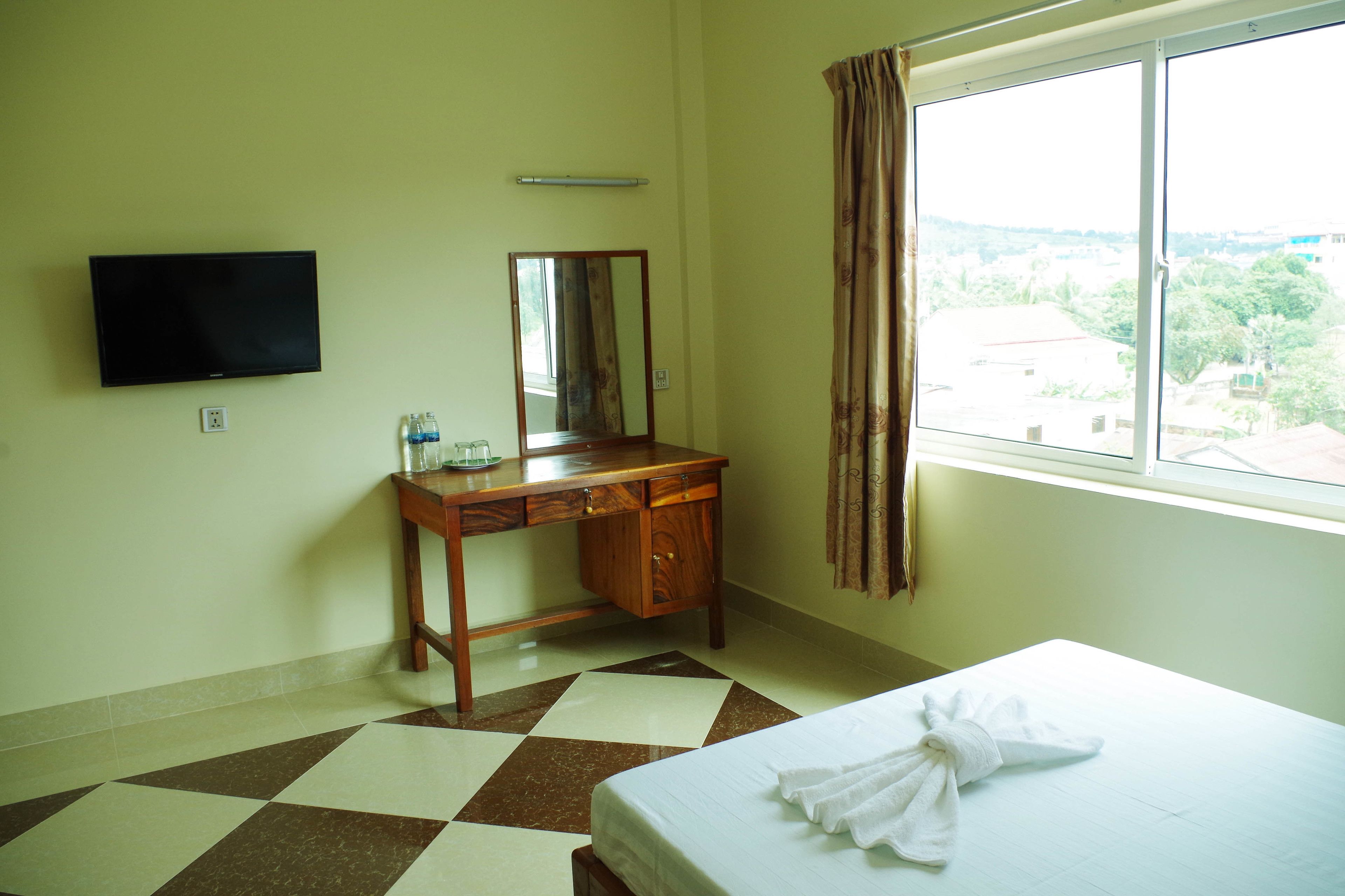 A hotel room with a bed, a wooden desk with a mirror, a television mounted on the wall, and a window with curtains.