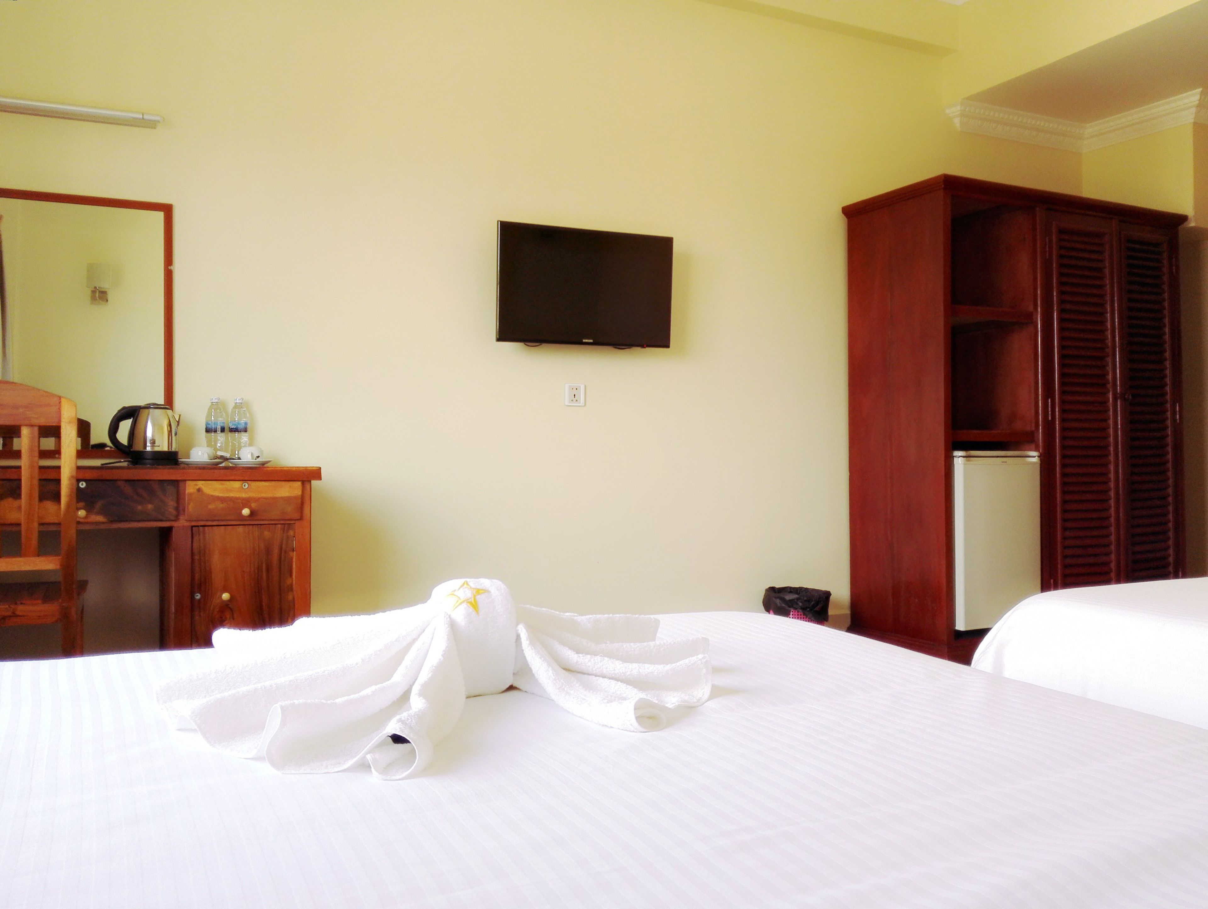 A hotel room with a bed, a desk with a kettle and bottles, a TV, and a wardrobe.