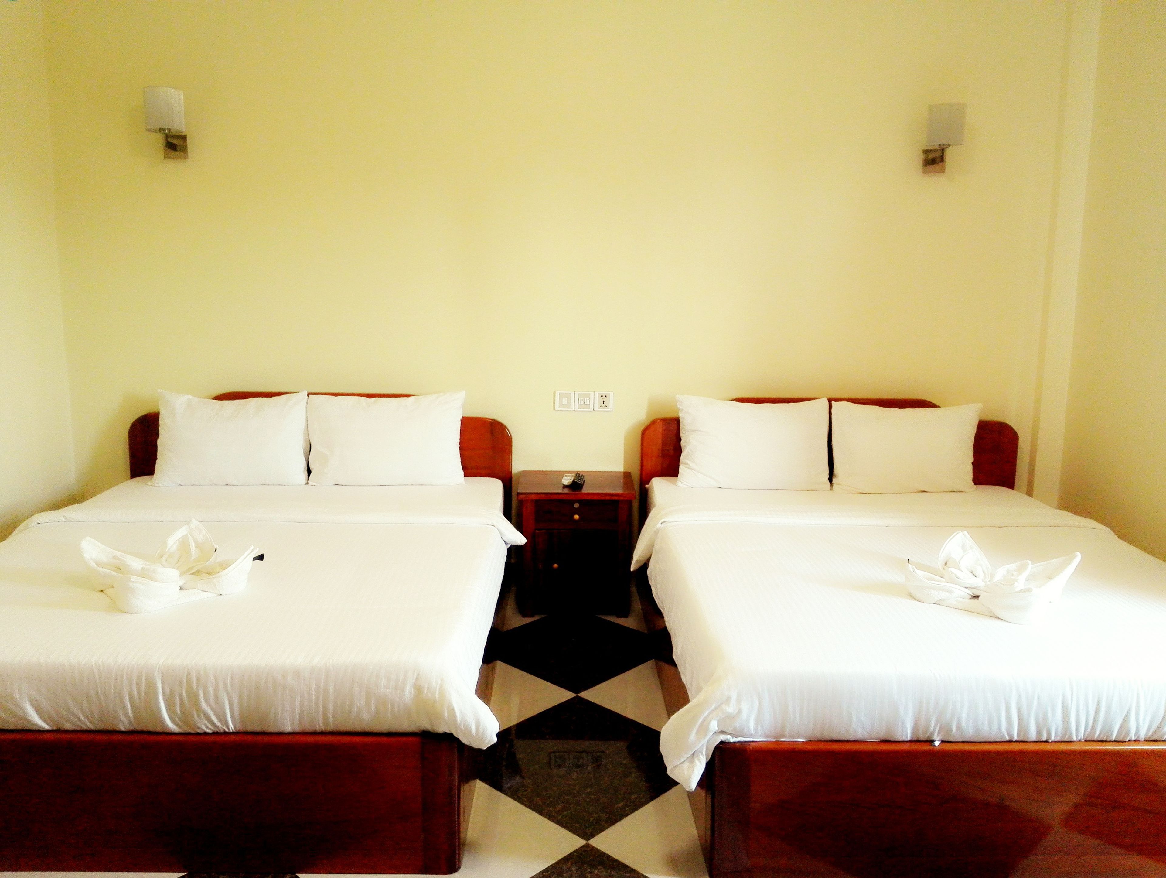 A hotel room with two beds, each with white linens and pillows, a small wooden nightstand, and a black and white checkered floor.