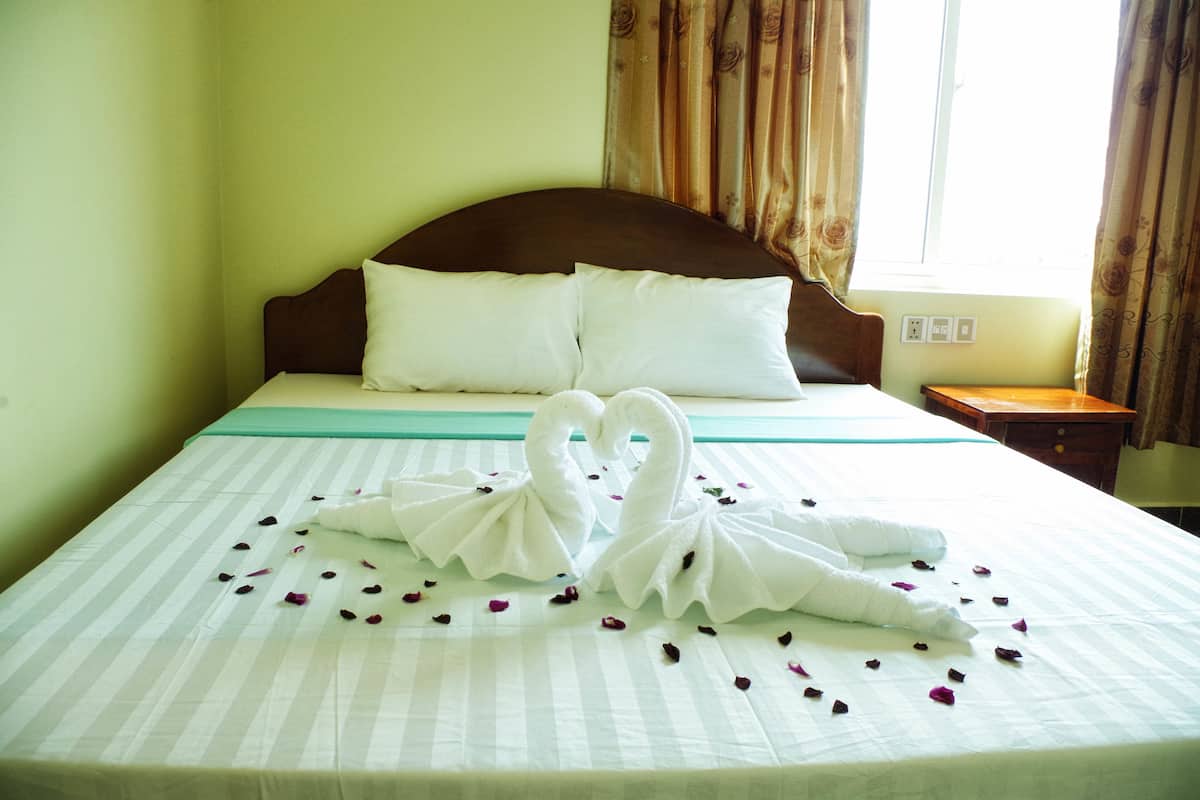A neatly made bed with a decorative swan towel arrangement and rose petals, a wooden headboard, and a window with curtains.