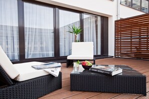 Un área de estar al aire libre moderna con un sofá de mimbre y un sillón blanco, una mesa de centro de vidrio con un frutero y una revista.