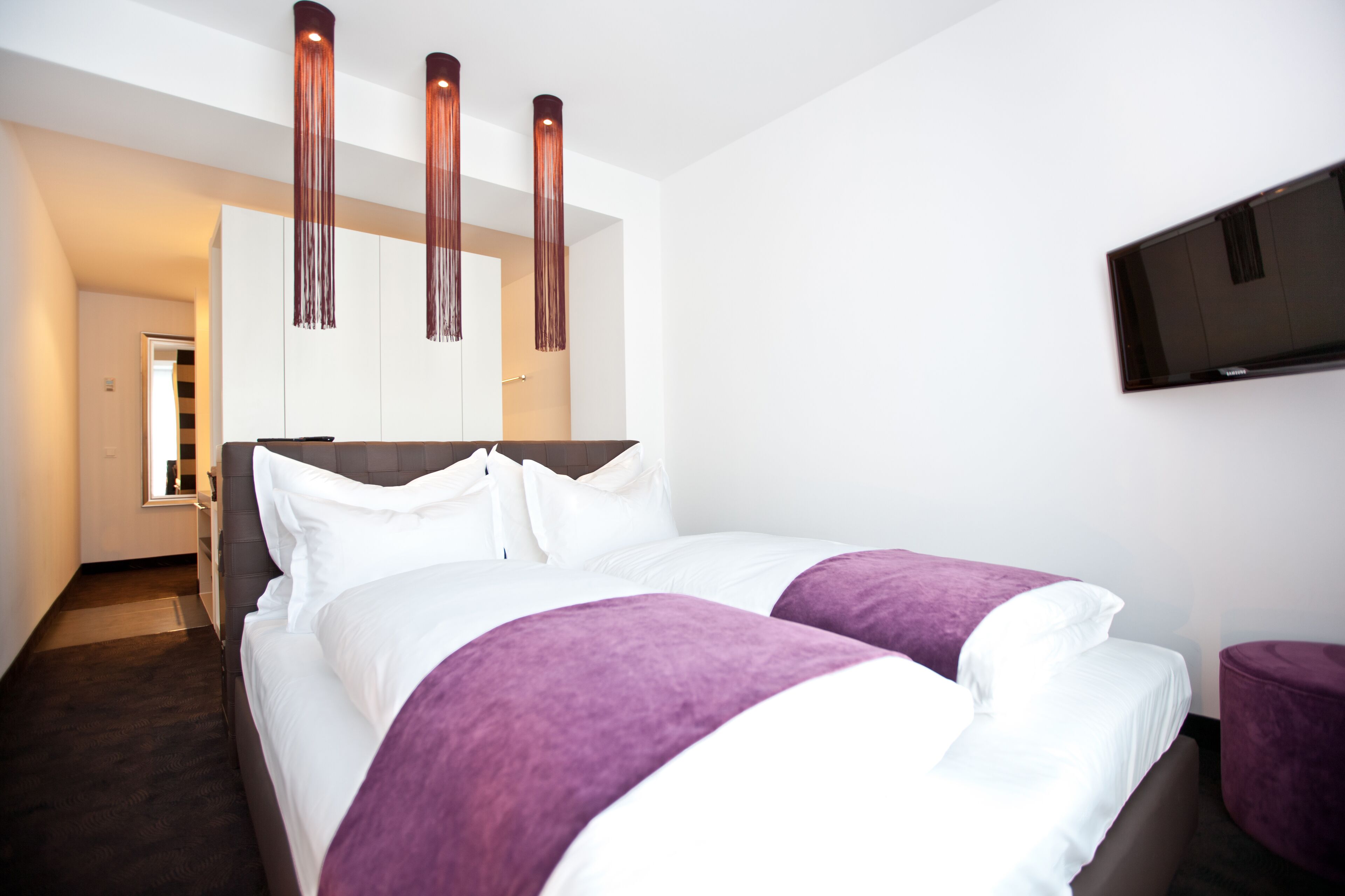 A modern hotel room with a large bed, a flat-screen TV mounted on the wall, and a hallway leading to another room.
