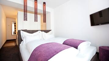 A modern hotel room with a large bed, a flat-screen TV mounted on the wall, and a hallway leading to another room.