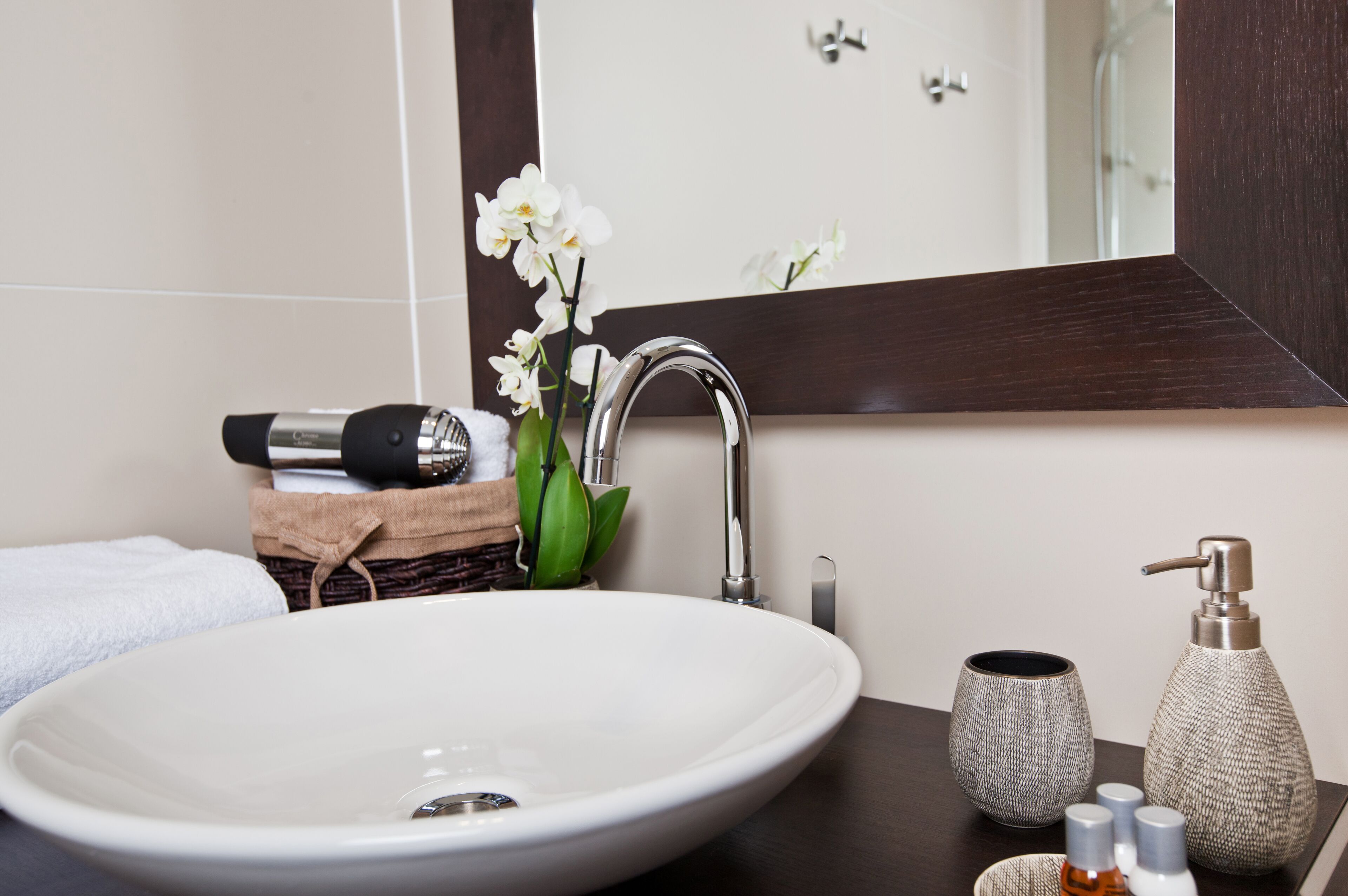 Een badkamer met een witte wastafel, een spiegel, een handdoek, een fles shampoo, een fles conditioner en een vaas met bloemen.