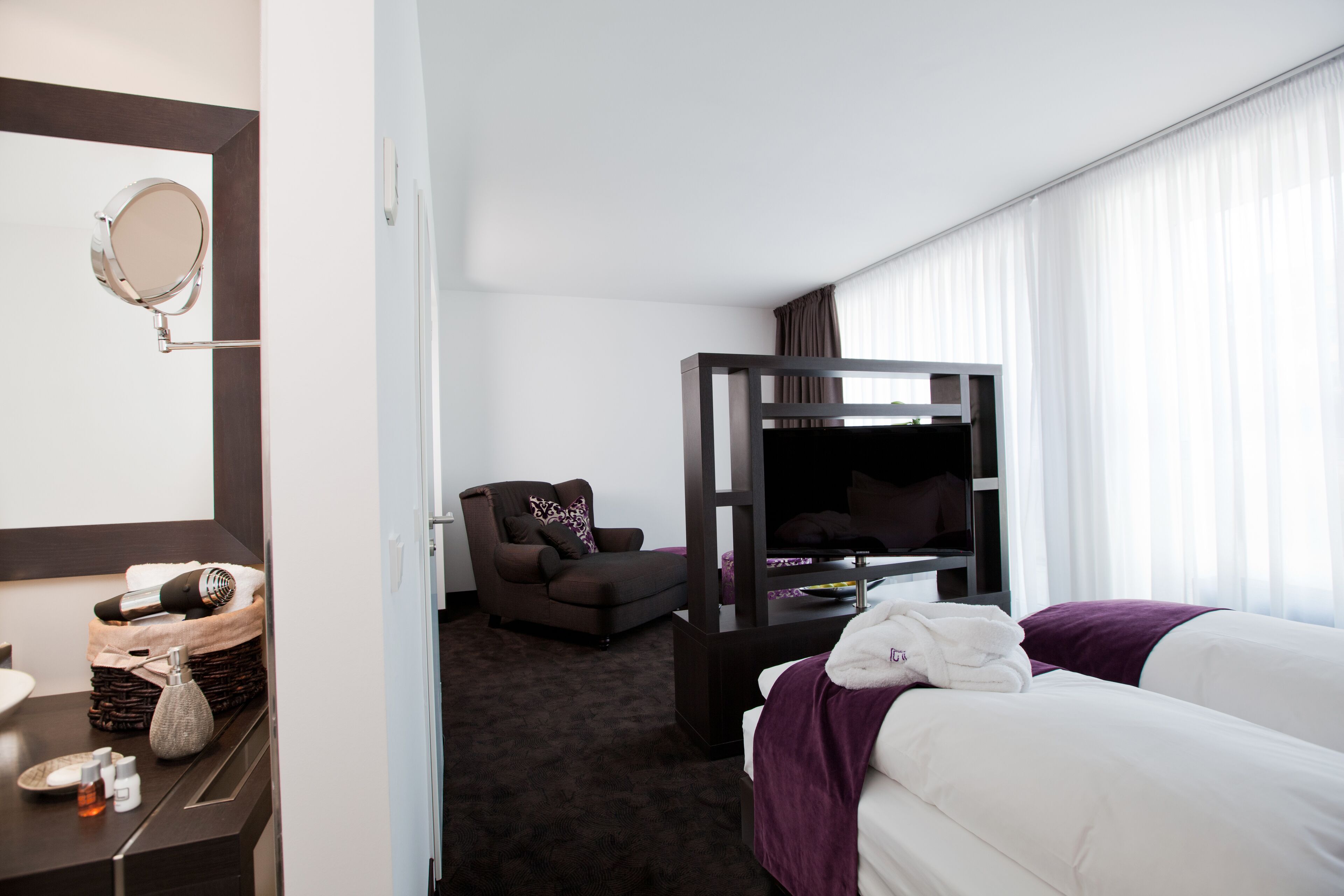 A hotel room with a bed, a TV, a chair, a vanity with a mirror, and a small table with toiletries.