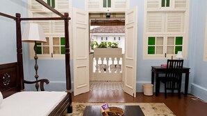 A bedroom with a four-poster bed, a bedside table with a lamp, a small table with a basket, and a view through an open door to a balcony with a railing. - PalanquInn Heritage Suites (George Town)