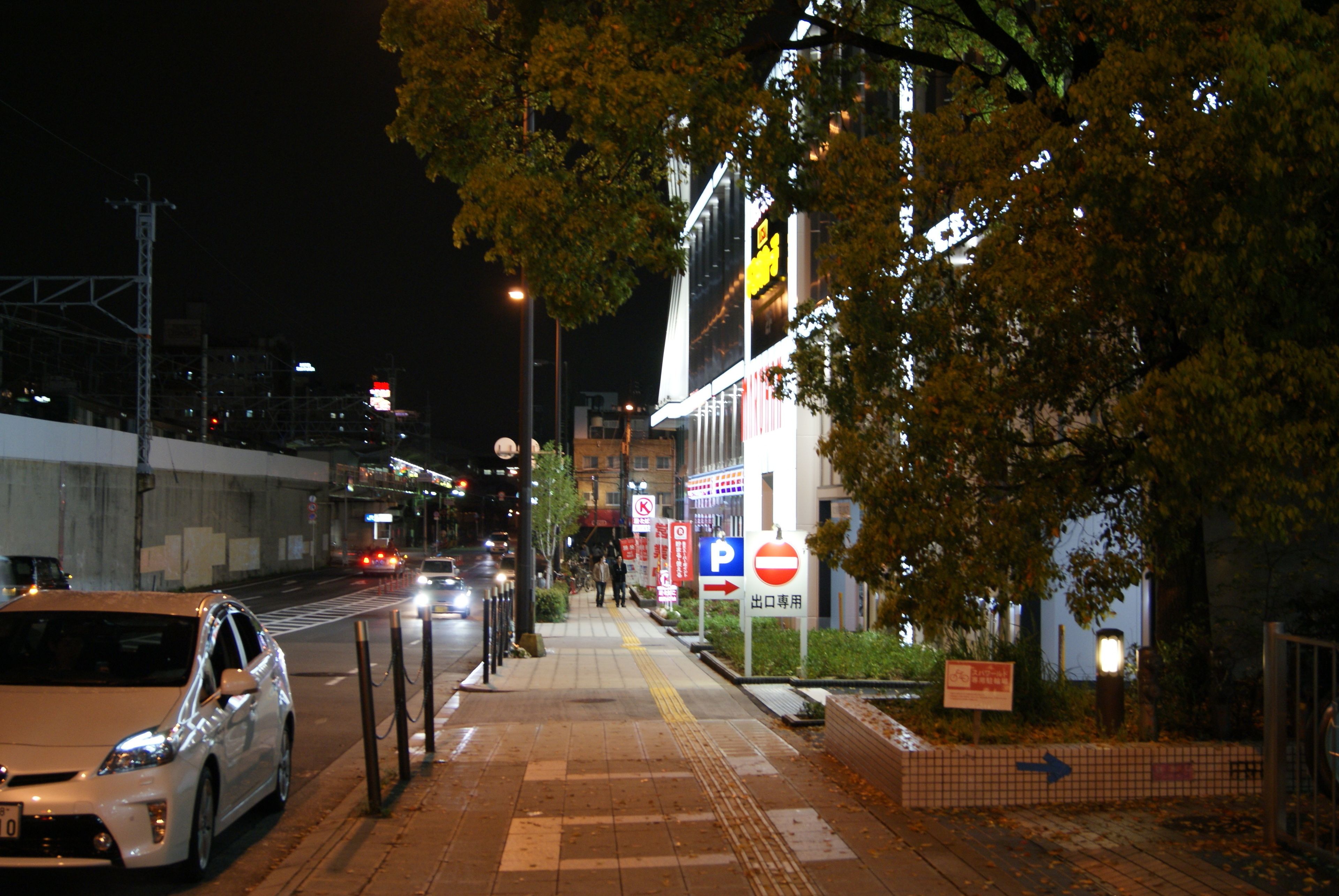 Scena notturna di una strada con auto parcheggiate sul lato, un attraversamento pedonale e un edificio con insegne illuminate.