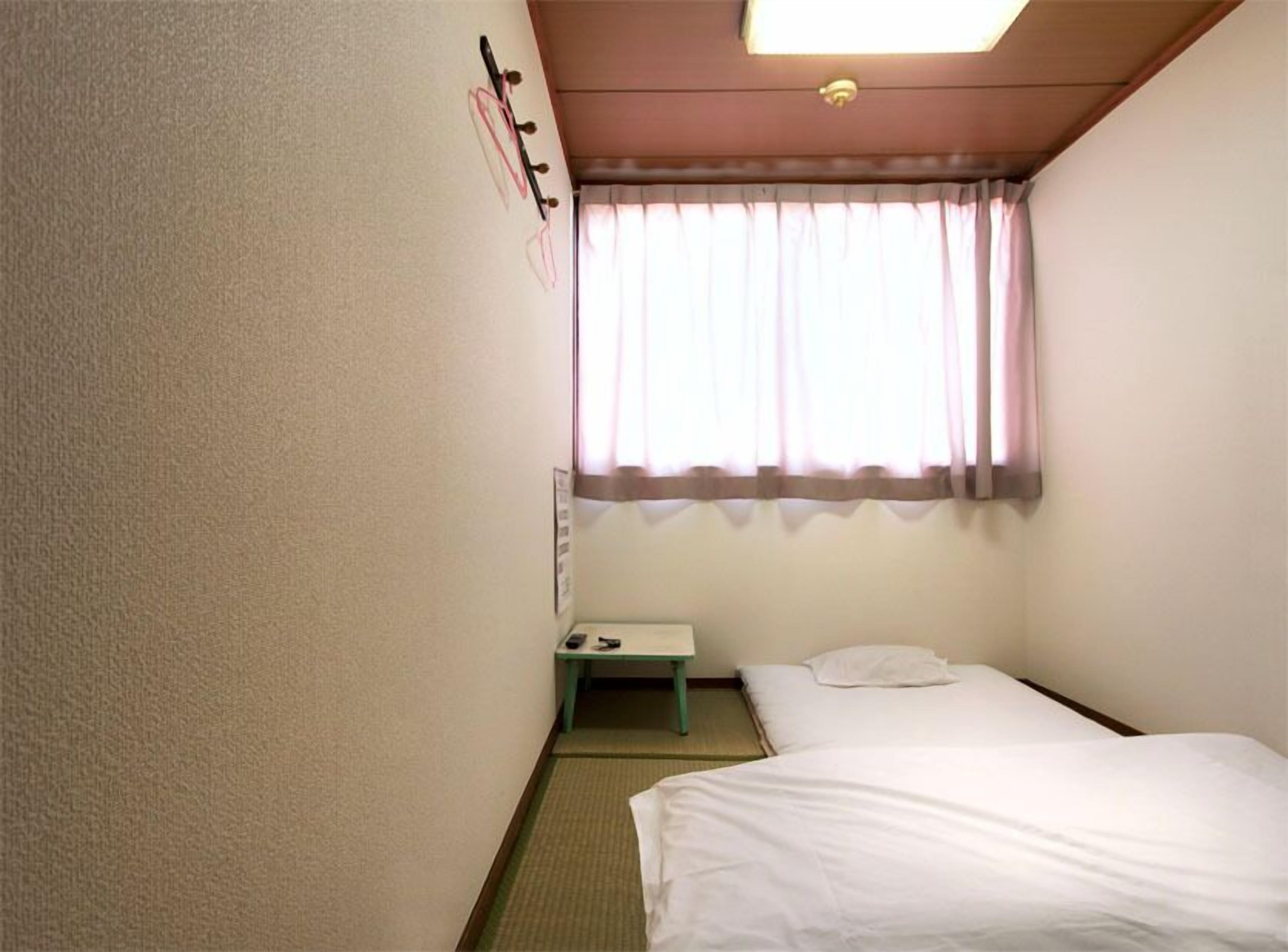 A small, single-bed room with a window, a bedside table, and a wall-mounted light fixture.