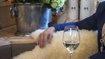 A room with a metal bucket of flowers, a fluffy white pillow, and a glass of white wine on a wooden table.