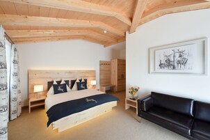 Un dormitorio con techo de madera, una cama con manta azul, un sofá de cuero negro, una mesita de noche con lámpara y un cuadro enmarcado en la pared.