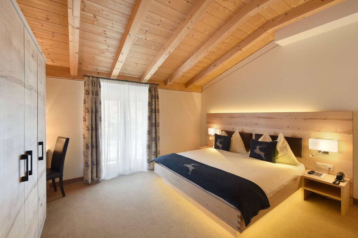 A bedroom with a wooden ceiling, a bed with a dark blue bedspread, a nightstand with a lamp, and a chair.