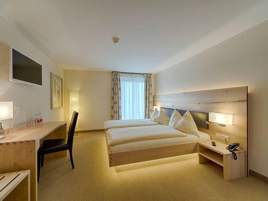 Habitación de hotel con cama, escritorio, silla, mesita de noche, lámpara y ventana con cortinas.