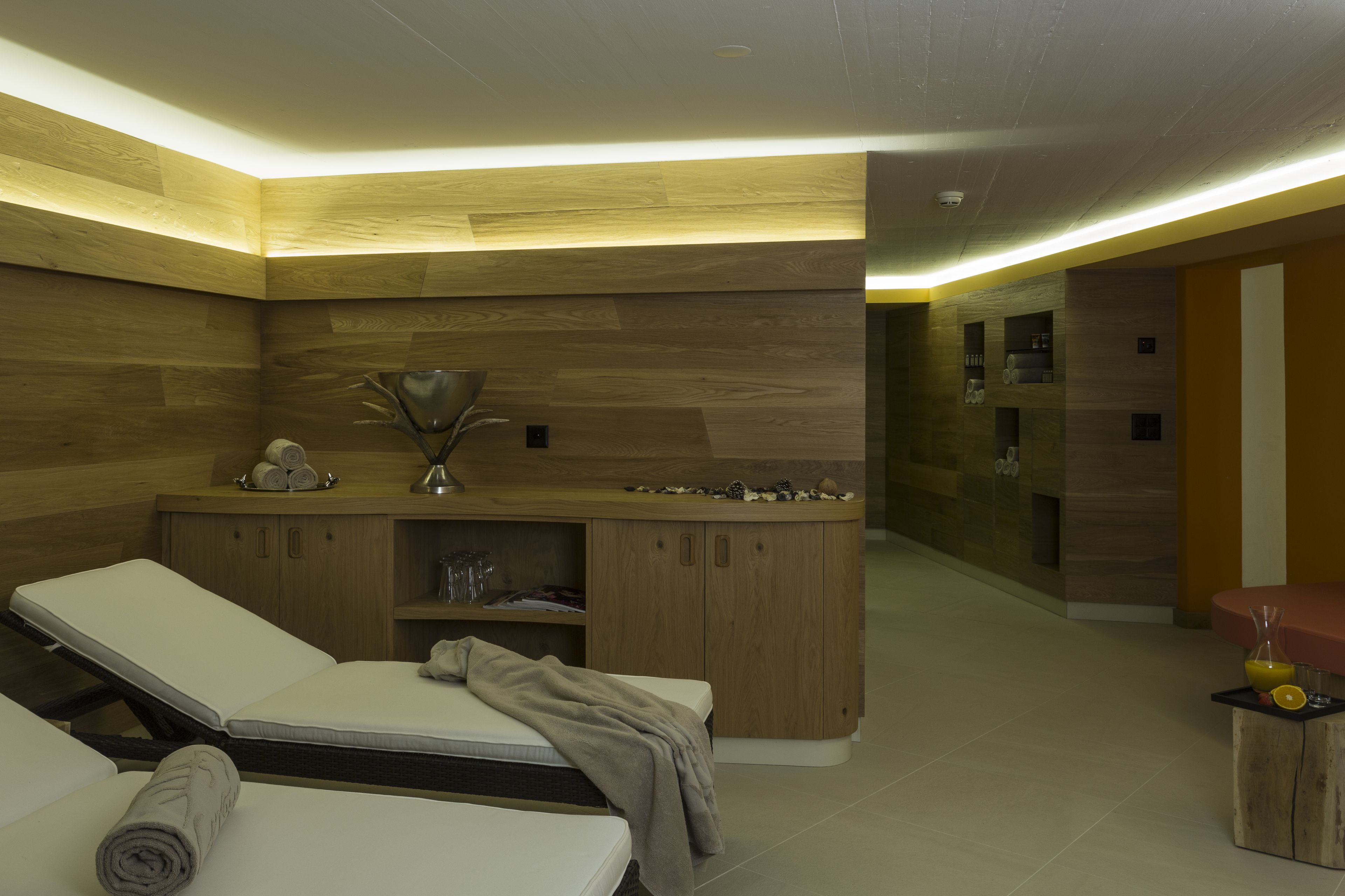 A modern spa room with wooden paneling, a massage table, a small wooden cabinet, and a seating area with a vase.