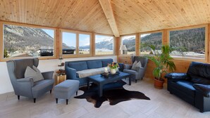 Amplia habitación con techo alto de madera, ventanas grandes con vistas a la montaña, un conjunto de sofá azul y un sillón de cuero.