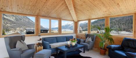 A spacious room with a high wooden ceiling, large windows offering mountain views, a blue sofa set, and a leather armchair.