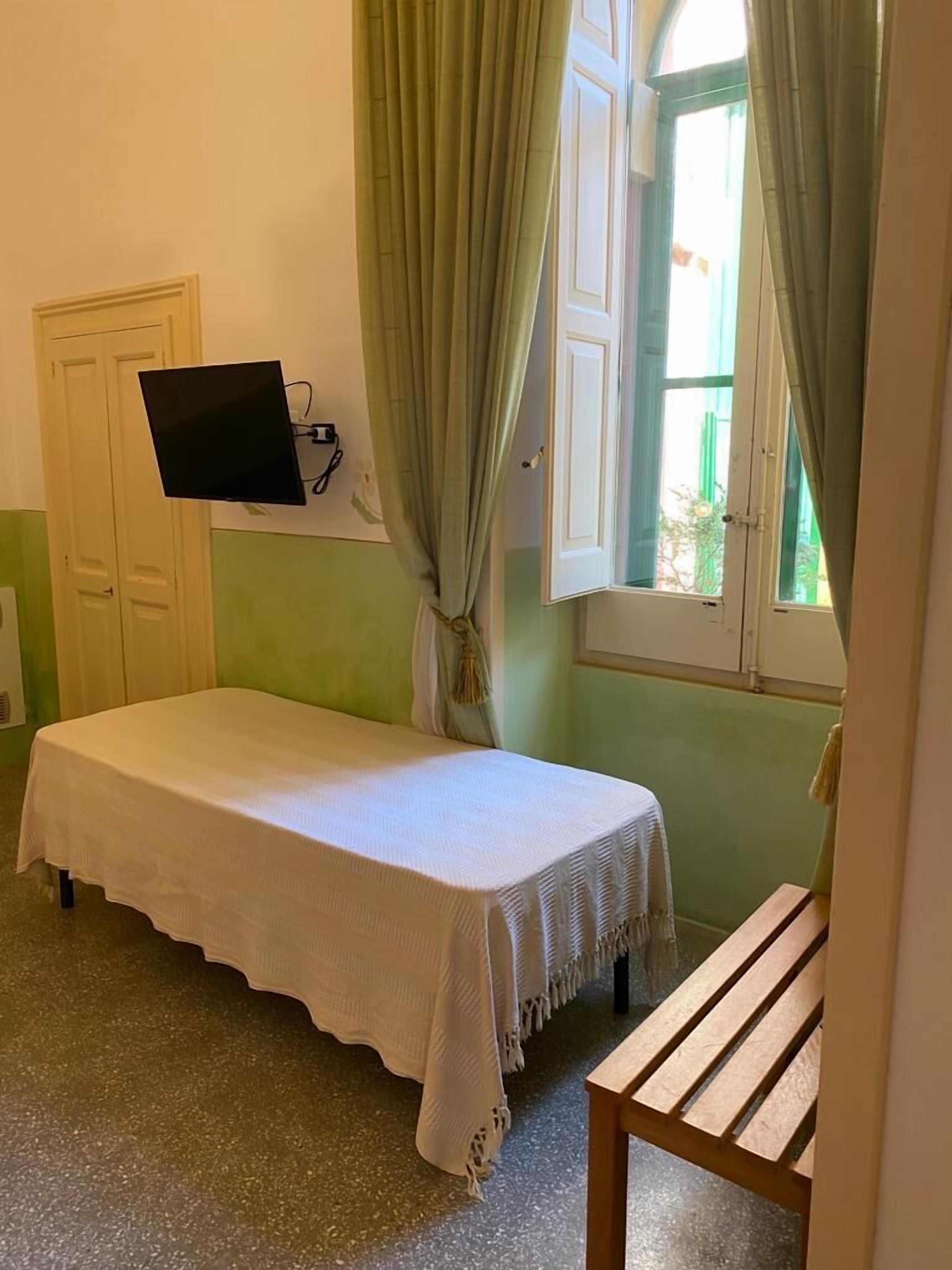 Une petite chambre avec un lit simple, un mur vert, une fenêtre avec des rideaux, un téléviseur fixé au mur et un banc en bois.