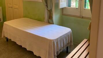 Une petite chambre avec un lit simple, un mur vert, une fenêtre avec des rideaux, un téléviseur fixé au mur et un banc en bois.