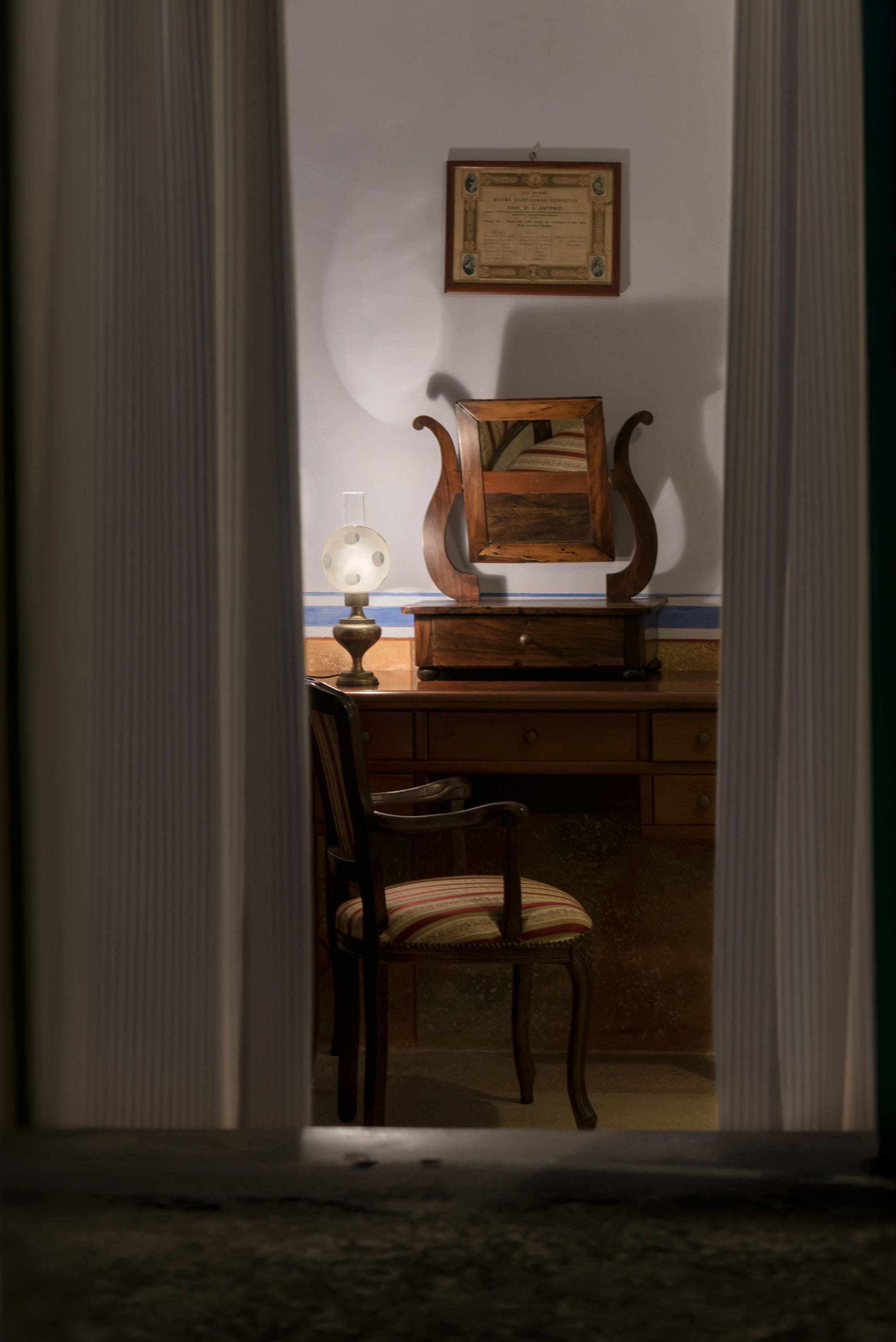 A room with a wooden desk, a chair, a lamp, and a framed certificate on the wall.