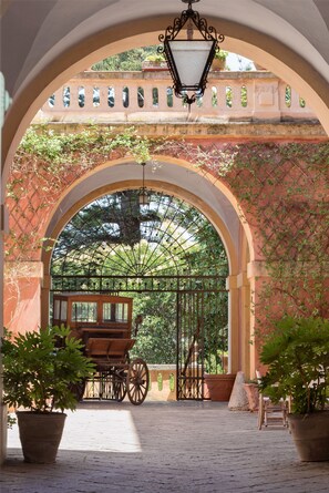 Une entrée en arc surmontée d’un portail en fer forgé, une calèche en bois et des plantes en pot.