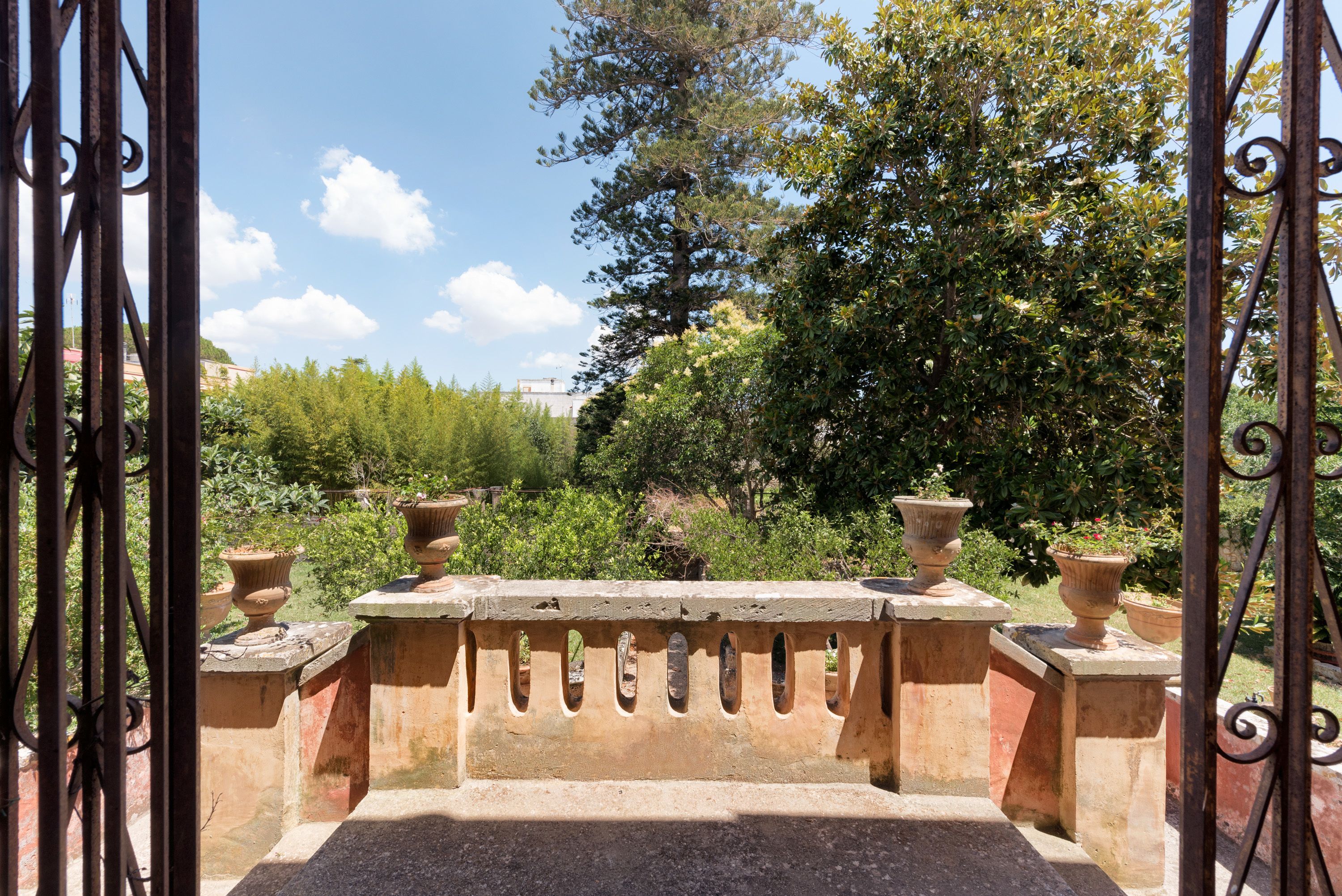 Ein schmiedeisernes Tor mit Steinbalustrade führt zu einem Garten mit Kübelpflanzen und Bäumen.