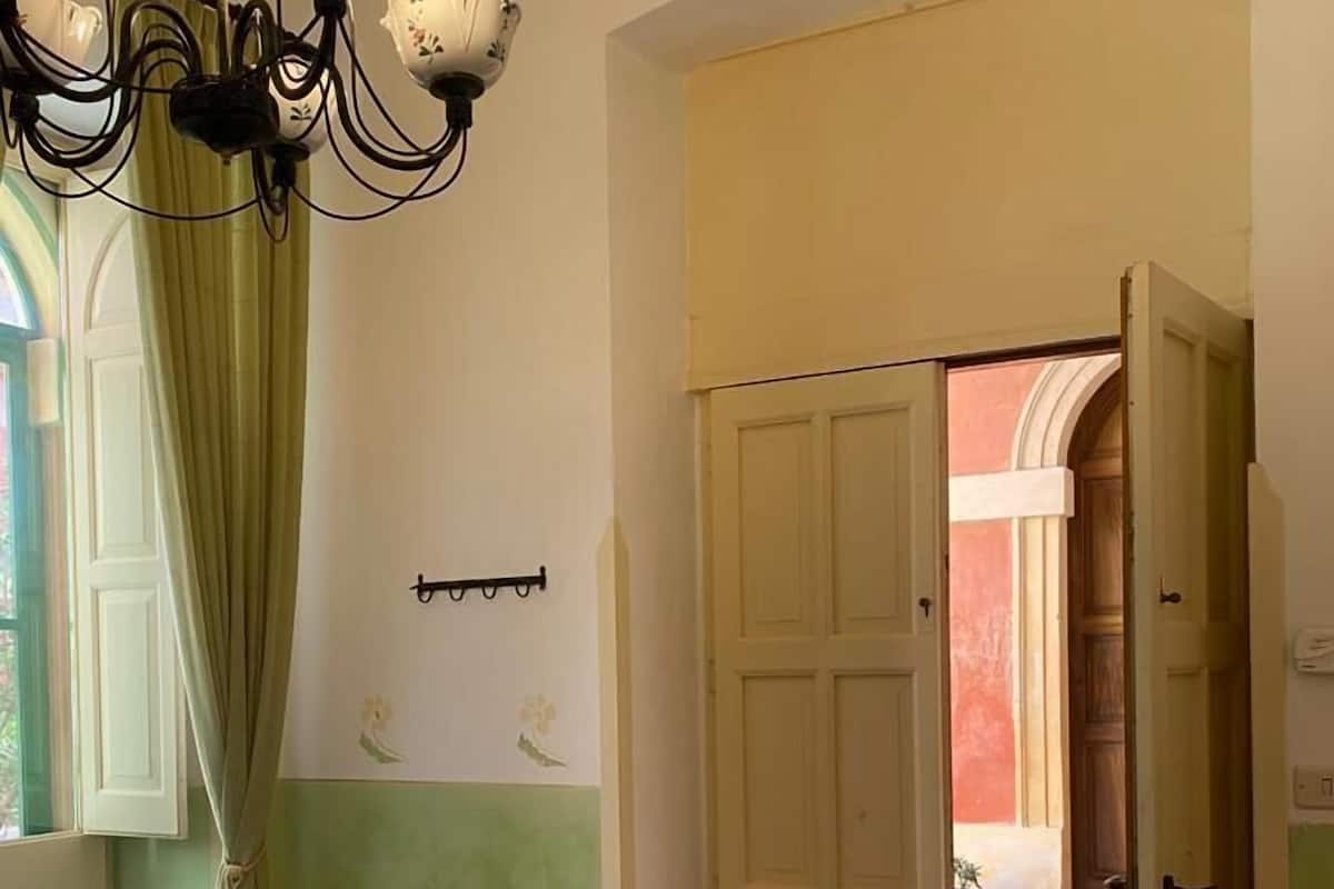 A hotel room with a bed, a small table, a bench, a chandelier, and a door leading to a courtyard.