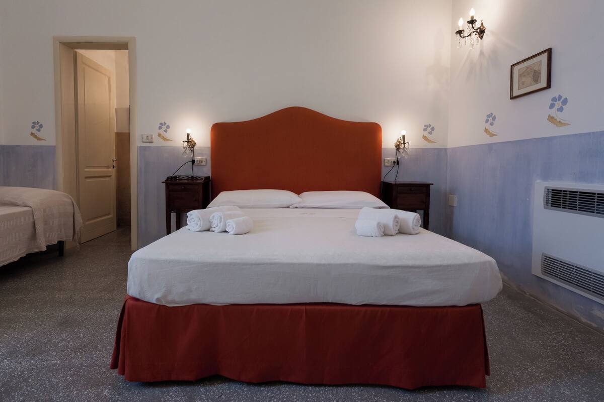 A neatly made bed with white linens and a red skirt, two bedside tables with lamps, a door to another room, and wall decorations.