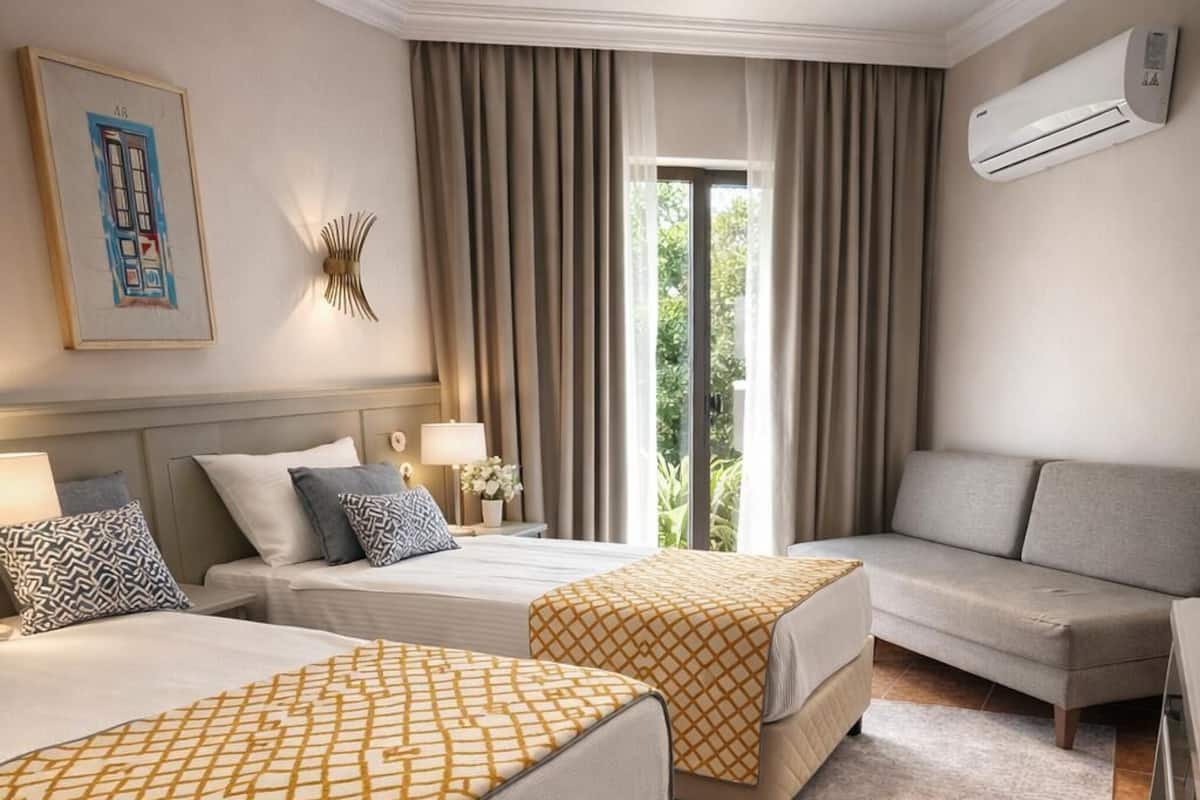 Ein Hotelzimmer mit zwei Betten, einem Sofa, einem Schreibtisch, einer Klimaanlage und einem Fenster mit Vorhängen.