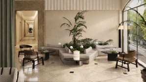 A spacious lobby with a modern design, featuring a central seating area with sofas and chairs, a large potted plant, and a view through a glass wall.