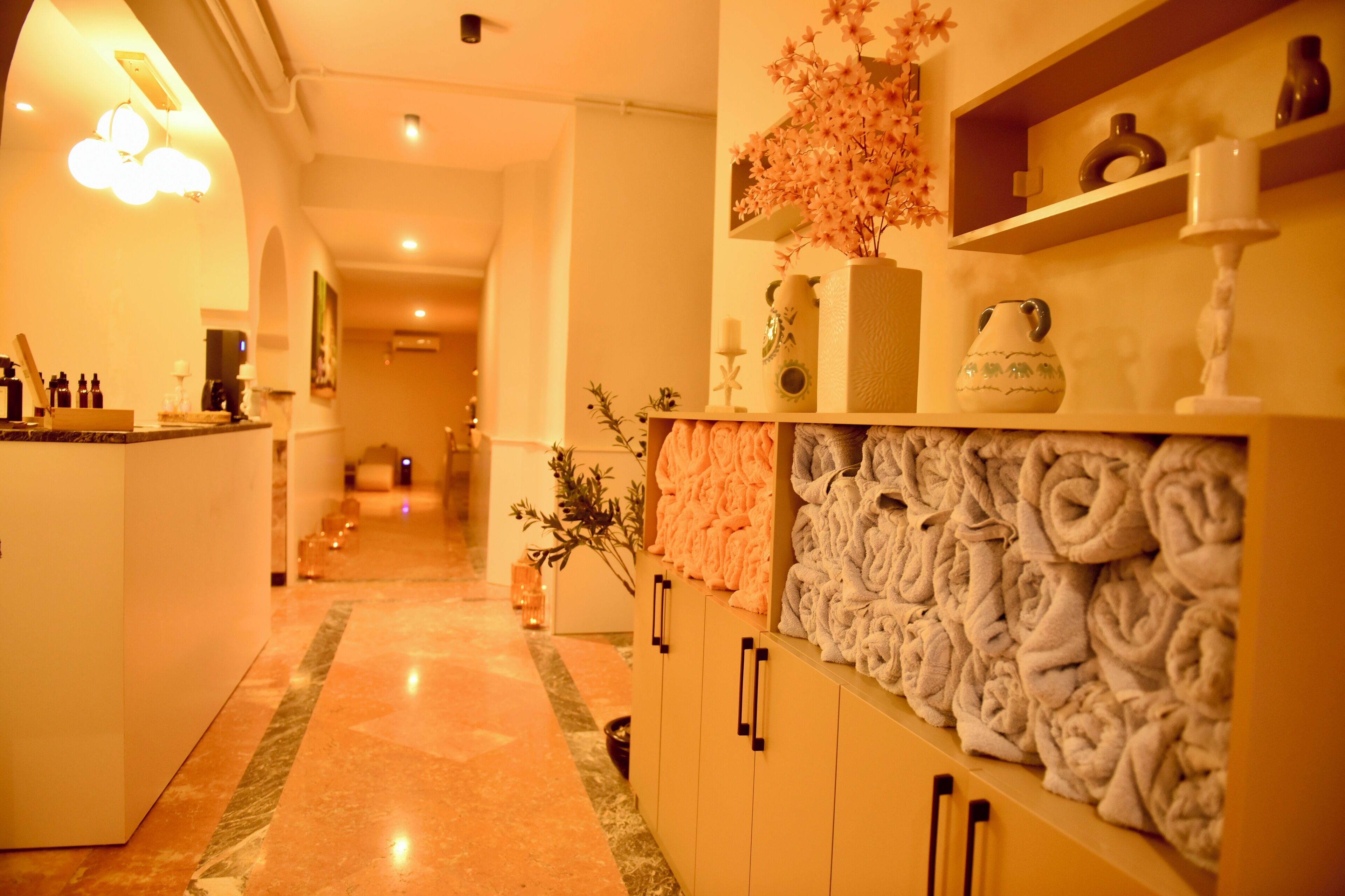 Un pasillo de hotel con piso de mármol, un mostrador de recepción, una pared con toallas enrolladas y una estantería con objetos decorativos.