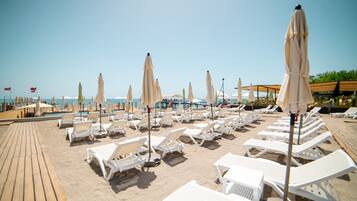 A beachside area with rows of white lounge chairs and beige umbrellas, a wooden deck, and a clear view of the sea.