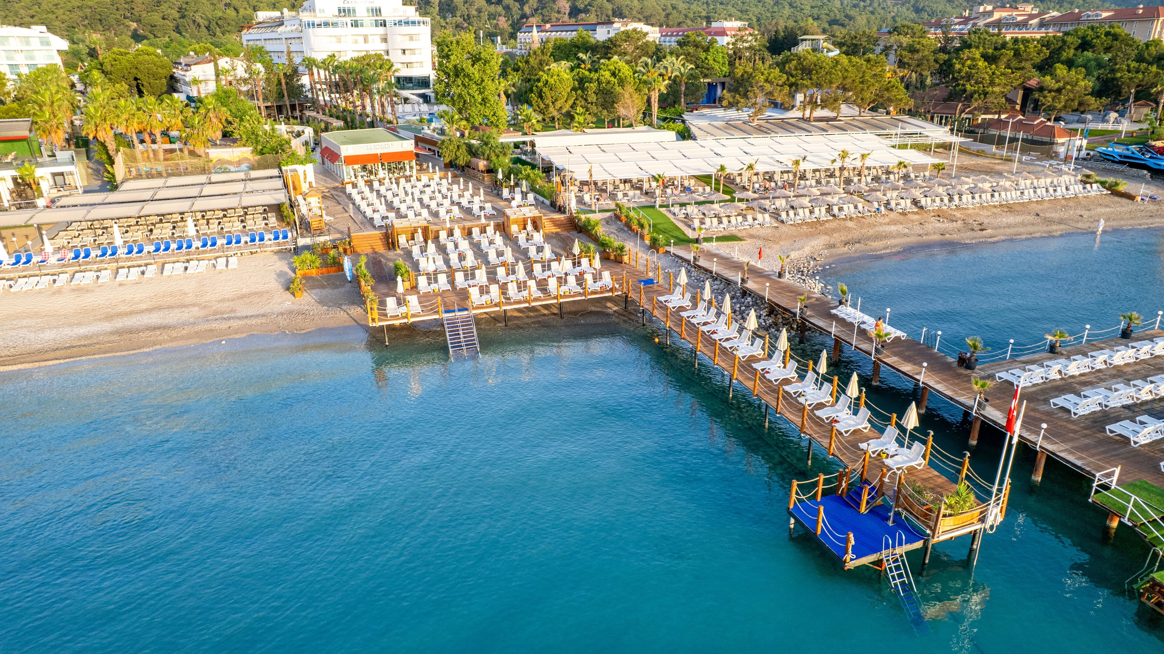 An aerial view of a waterfront hotel with multiple levels of sun loungers and umbrellas, a dock extending into the water, and a backdrop of buildings and greenery.