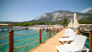 A wooden pier with white lounge chairs and umbrellas, leading to a beach with clear turquoise water and a mountain in the background. - Rose Garden Premium Hotel - All inclusive (Kemer)
